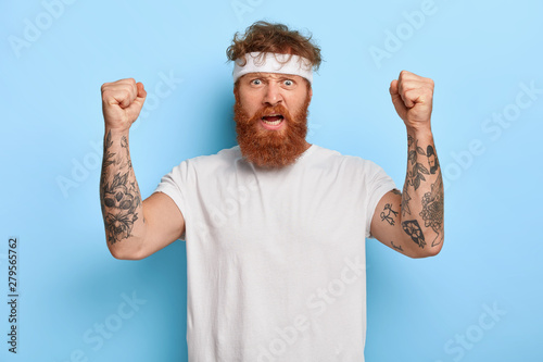 Strong angry sportsman with red hair, raises tattooed arms, shows his power, exclaims with dissatisfaction, wears white headband and t shirt, isolated on blue background. Sport and strength.