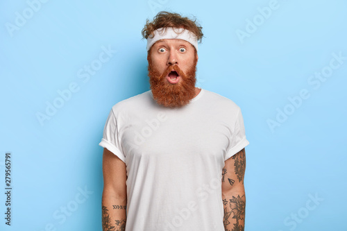 Obraz na płótnie Indoor shot of surprised bearded man with red hair, stares with astonishment, has gymnastic every day, dressed in white casual clothes, headband, models over blue wall