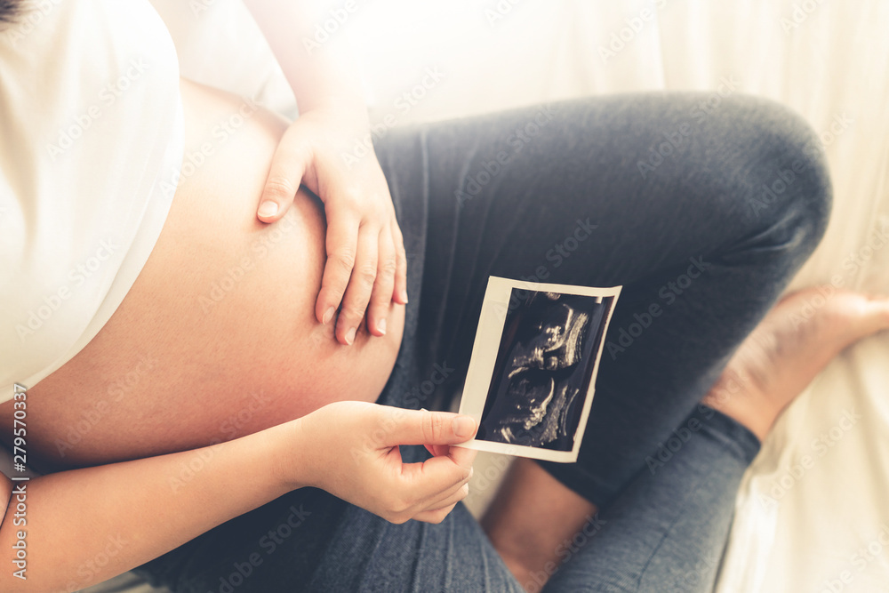 Pregnant woman feeling happy at home while taking care of her child. The young expecting mother holding baby in pregnant belly. Maternity prenatal care and woman pregnancy concept. - obrazy, fototapety, plakaty 