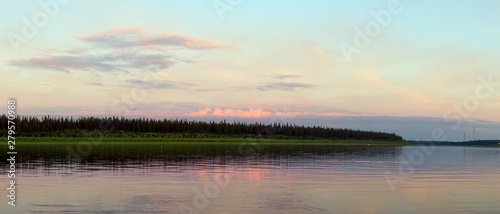 Bright orange sunset behind the clouds of sun on the banks of the Northern river Viluy under the blue sky in the spruce forest and silhouettes of boats and people in Yakutia.