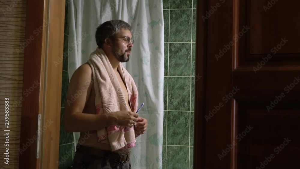 Adult man with a beard brushes his teeth in the bathroom. Excess weight. Indoors. Towel on the neck. Green dark tile. Dim lighting. View of the belt. Adult man with a beard brushes his teeth in the bathroom. Excess weight. Indoors. Towel on the neck. Green dark tile. Dim lighting. View of the belt.