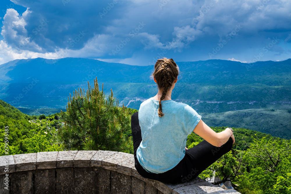 Montenegro, Beautiful young girl sitting in tailor seat on a stone wall enjoying wide view over green nature landscape bjelopavlici plain lowlands valley from mountains near ostrog monastery