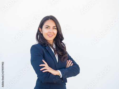 Happy confident female business leader posing in studio background. Beautiful young Latin woman in formal suit standing with arms folded and smiling at camera. Successful businesswoman concept photo