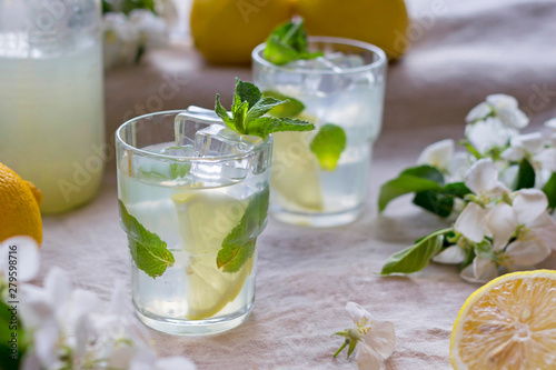 Bottle and glasses of cold lemon drink, ice, mint leaves, slices, white apple flowers on linen tablecloth. Close-up, copy space