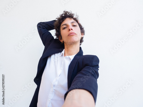 Confident androgynous young woman holding cell phone in hand and taking selfie. Self portrait of female professional in office suit. Poser concept © Mangostar