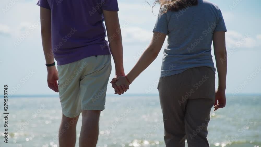 Man and Woman Standing on the Coast of the Sparkling Sea in Windy Summer Day. Young Couple Holding Hands and Looking at the the Sea. Slow Motion. Lifestyle, Love, Dating, Vacation Concept Man and Woman Standing on the Coast of the Sparkling Sea in Windy Summer Day. Young Couple Holding Hands and Looking at the the Sea. Slow Motion. Lifestyle, Love, Dating, Vacation Concept
