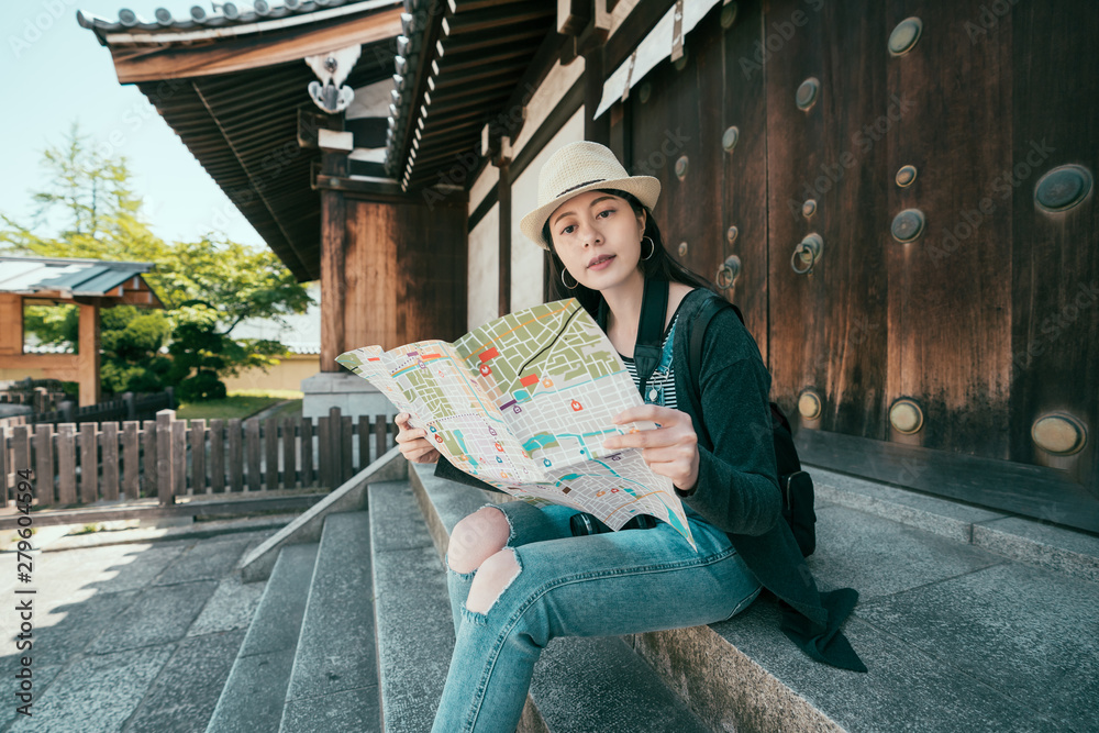Obraz premium Young woman traveler with backpack and hat looking paper map in shitennoji temple osaka japan. Traveling asia in summer holidays. relaxed girl visitor in shrine sitting on rock stairs thinking way.