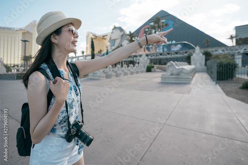 pyramid and sphinx in Las Vegas Nevada. cheerful young girl backpacker carrying vintage camera searching direction of tourism attraction. side view of excited asian woman traveler point finger away photo