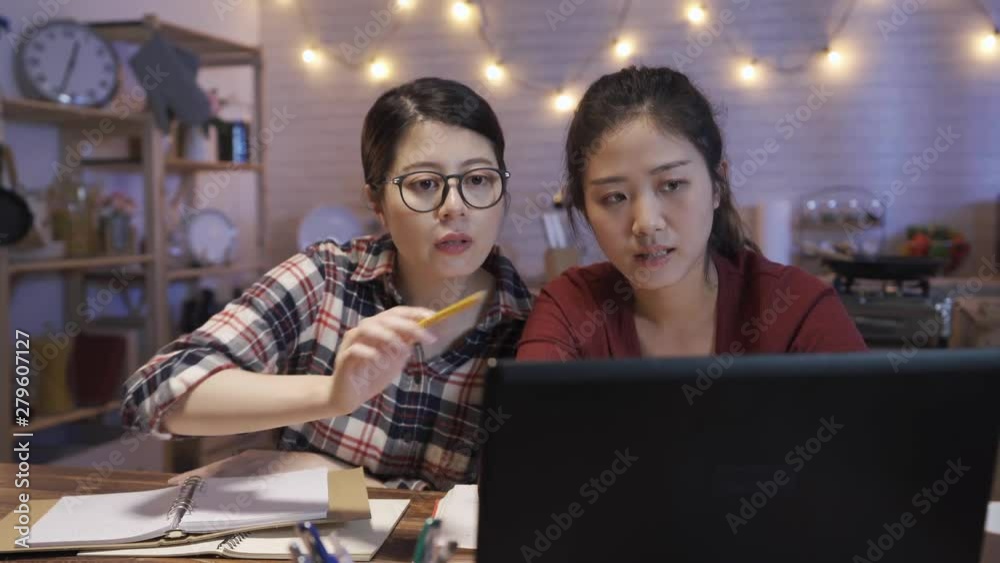 Cheerful international female students talking to each other while doing homework task in night home kitchen. two young college girls having discussion on laptop computer in dark dining room in house Cheerful international female students talking to each other while doing homework task in night home kitchen. two young college girls having discussion on laptop computer in dark dining room in house