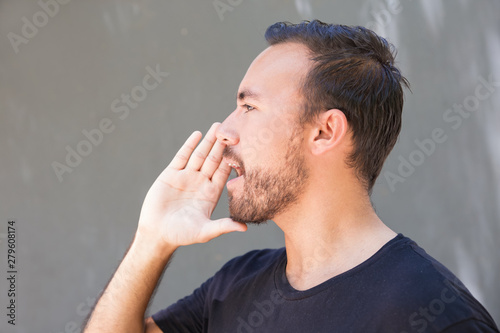 Young man screaming and looking aside. Handsome bearded young man yelling and looking away on grey background. Gesture concept