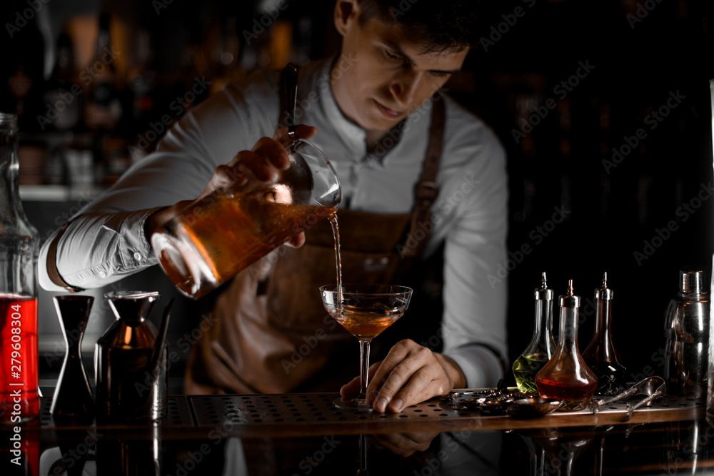 Bartender pouring a brown alcoholic cocktail from the measuring cup to the glass in the dark