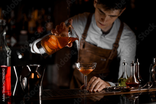 Attractive male bartender pouring a brown alcoholic cocktail from the measuring cup to the glass in the dark