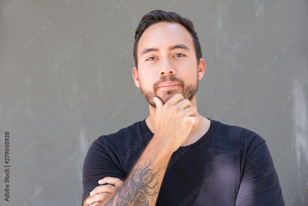Bearded man standing with hand on chin. Handsome tattooed man in black t-shirt standing with hand on chin and looking at camera on grey background. Gesture concept Bearded man standing with hand on chin. Handsome tattooed man in black t-shirt standing with hand on chin and looking at camera on grey background. Gesture concept