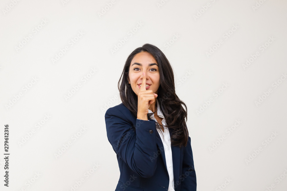 Young businesswoman gesturing for silence. Happy young woman holding finger on lips and smiling at camera isolated on grey background. Gesture concept Young businesswoman gesturing for silence. Happy young woman holding finger on lips and smiling at camera isolated on grey background. Gesture concept