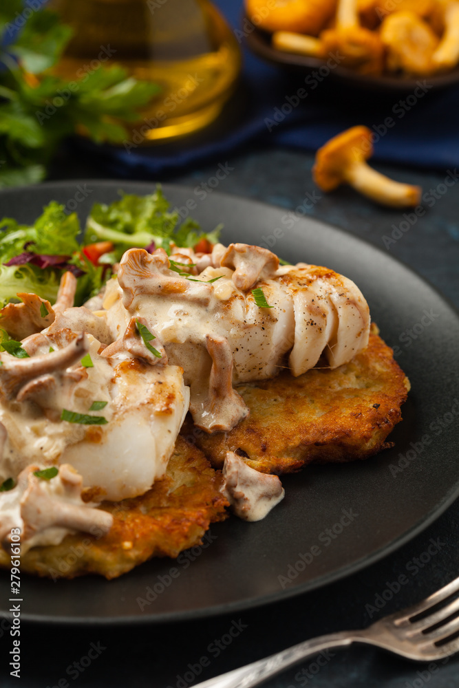Fried cod, served on potato pancakes with mushrooms sauce and salads. Portion on a black plate. Dark background. Fried cod, served on potato pancakes with mushrooms sauce and salads. Portion on a black plate. Dark background.