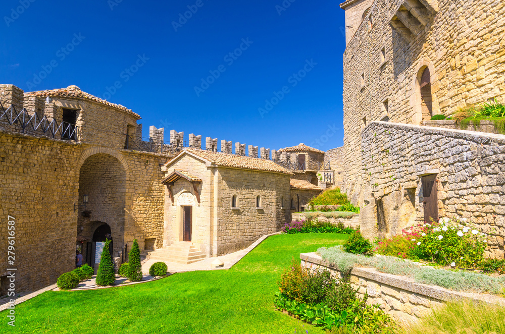 Courtyard with green grass lawn of Prima Torre Guaita first medieval tower with stone brick fortress wall with merlons on Mount Titano rock, blue sky white clouds background, Republic San Marino