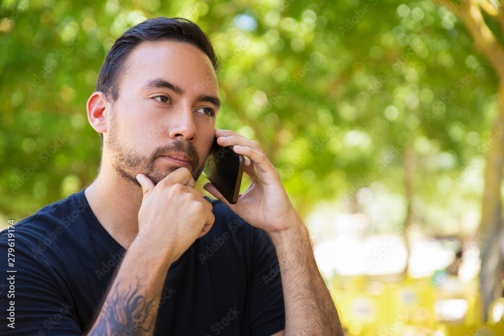 Serious young man talking by smartphone. Handsome bearded man in black t-shirt talking by cell phone and looking away outdoor. Communication concept Serious young man talking by smartphone. Handsome bearded man in black t-shirt talking by cell phone and looking away outdoor. Communication concept