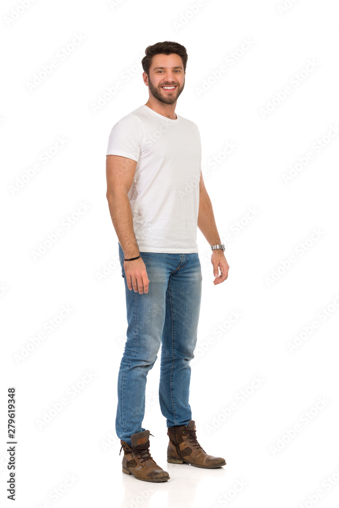 Smiling Young Man In Jeans And White T-shirt Is Standing And Looking At Camera. Smiling Young Man In Jeans And White T-shirt Is Standing And Looking At Camera.