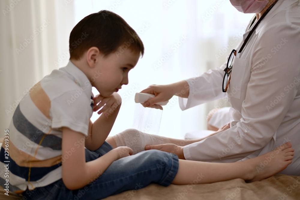 The hands of the doctor carefully bandage the knee of the child who is distressed from the injury. The hands of the doctor carefully bandage the knee of the child who is distressed from the injury.