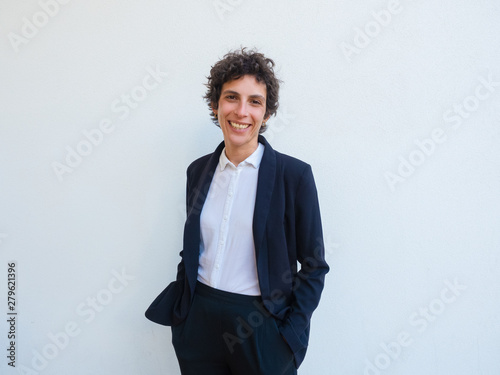 Happy androgynous businesswoman posing in studio. Middle aged woman in office suit standing over white background, keeping hands in trousers pocket and smiling at camera. Business portrait concept photo