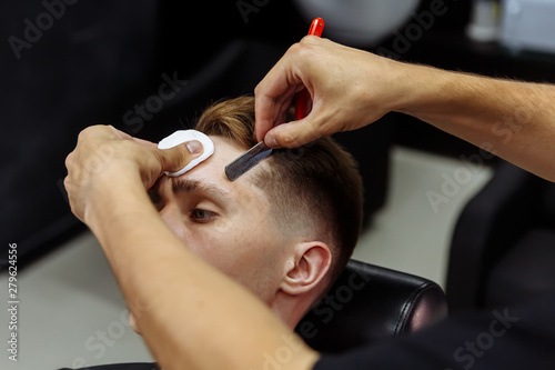 Professional barber with open cutthroat razor shaves hair on man's temples. Attractive male is getting a modern haircut in barber shop. Close up.