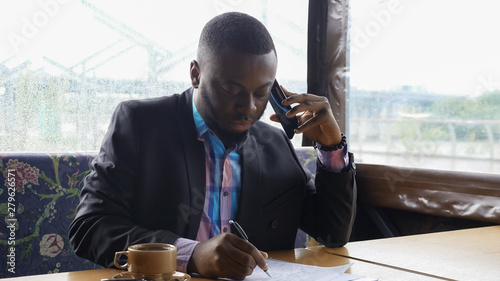 Black guy is talking on smartphone. Afro american businessman is calling on mobile phone sitting in summer tent cafe. He is drinking a cup of coffee at table. He wears shirt and suit jacket. photo