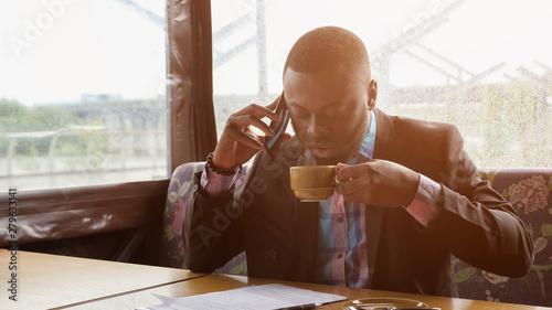 Black guy is talking on smartphone. Afro american businessman is calling on mobile phone sitting in summer tent cafe. He is drinking a cup of coffee at table, sunlight. He wears shirt and suit jacket. photo