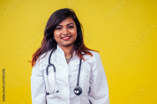 Female indian young and beautiful blond woman gynecologist doctor using stethoscope in a white medical coat on a yellow background in the studio
