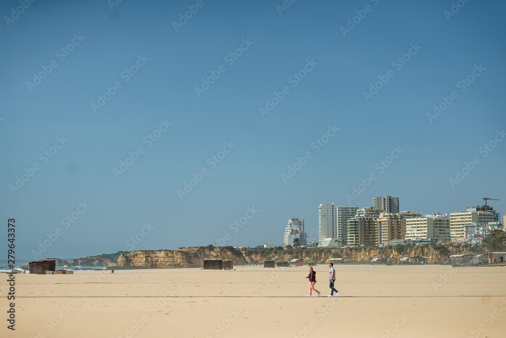 Happy young couple enjoying sunny days on the beach. Man and woman playing with sand. Love story near the ocean. Smiling attractive pair embracing and holding hands on the background of buildings. Happy young couple enjoying sunny days on the beach. Man and woman playing with sand. Love story near the ocean. Smiling attractive pair embracing and holding hands on the background of buildings.
