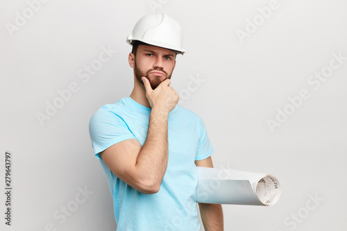 Pensive man with hand on his chin holding blueprint looking at the camera .puzzled man in hat. serious guy making up idea, plan. man solving a problem . isolated hwite background photo