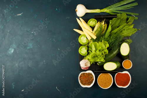 THE VEGETABLES IN THE BOX ON THE STONE A DARK BACKGROUND. YOUNG GREENS ONION GARLIC ZUCCHINI BRIGHT SPICES LAY IN A WOODEN TRAY WITH HANDLES ON A DARK TEXTURED BACKGROUND.