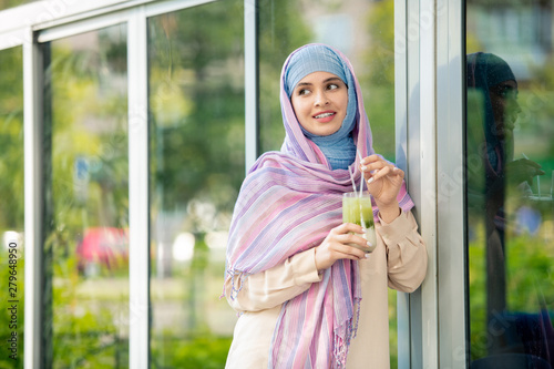 Young female in muslim hijab having refreshing drink outdoors