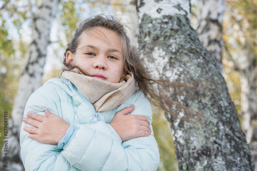 Frozen girl in scarf and blue down jacket hugging herself with hands from the cold near the birch in an autumn cool day.