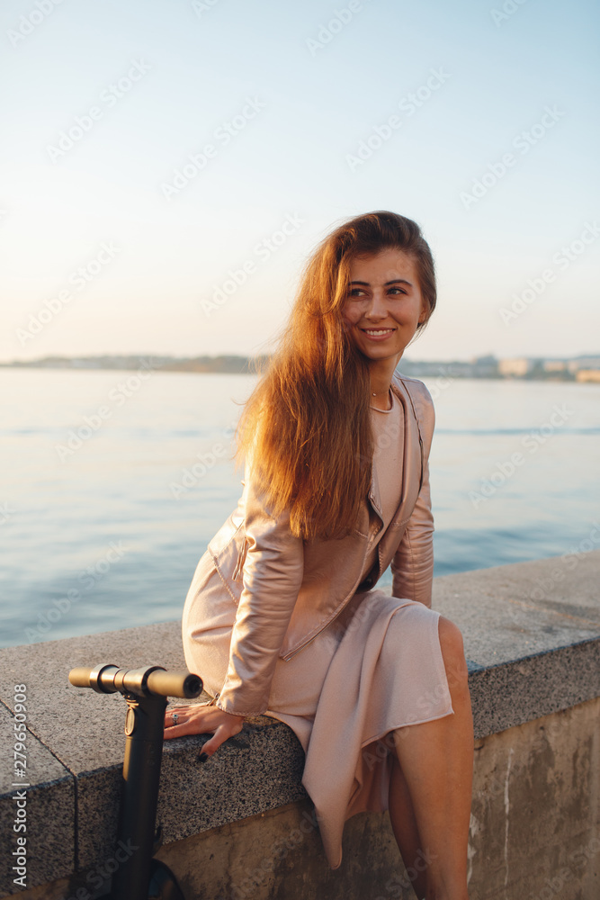 Portrait of a smiling pretty girl in pink clothes, rides on a  scooter and looks at the camera, background city in the sunset. Walk on a kick scooter.Beautiful woman riding a scooter, walking, leisure