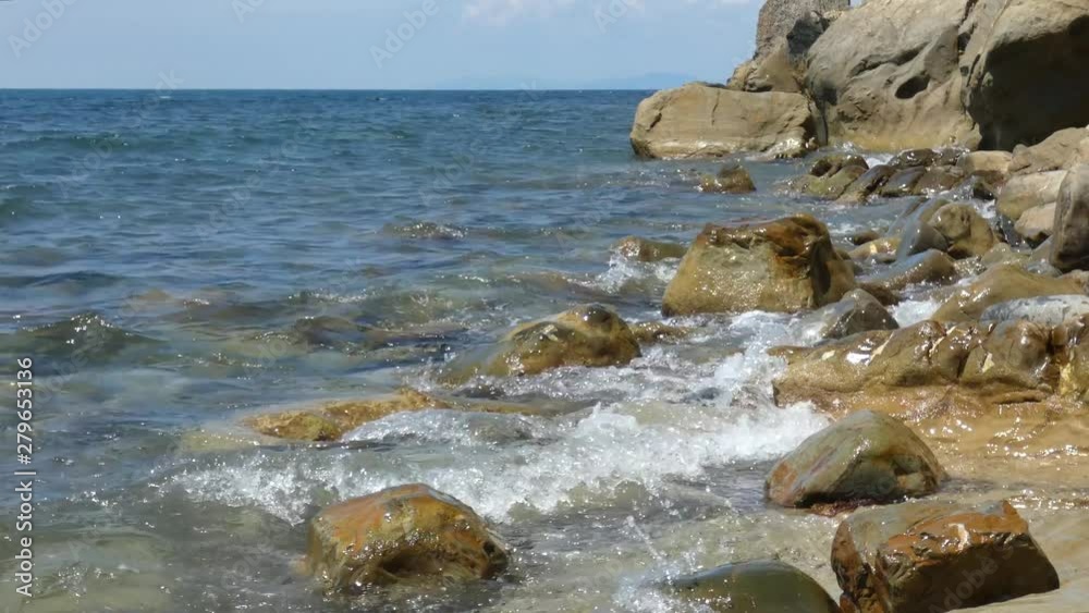 Sea surf on big rocks. Water waves on beach with multicolored polished stones. Open sea with horizon line at the back. Sunny summer day. Natural background. Sea surf on big rocks. Water waves on beach with multicolored polished stones. Open sea with horizon line at the back. Sunny summer day. Natural background.