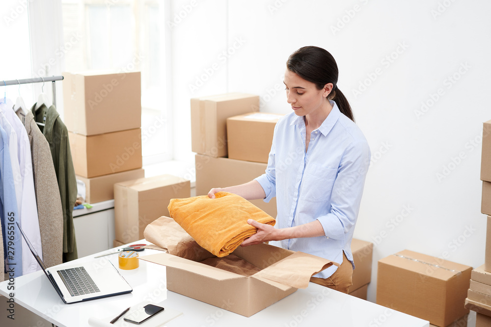 Young female online shop manager putting folded velvet yellow trousers into box