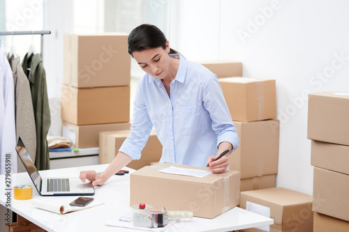 Young female manager of online shop writing down address of client on top of box