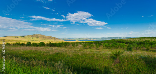 Large panorama of blue sky with white clouds  fields and meadows with green grass  against the mountains. Composition of nature. Rural summer landscape.