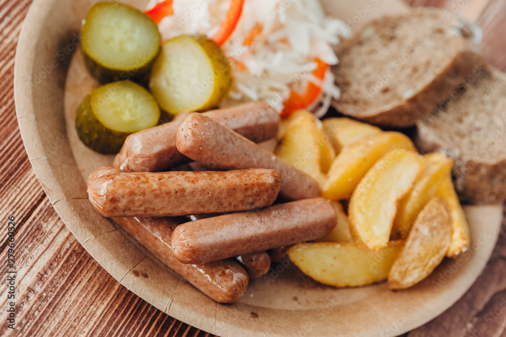 Tasty sausages with fried potatoes, cucumber, cabbage and chlobe in a paper plate on a wooden table.
