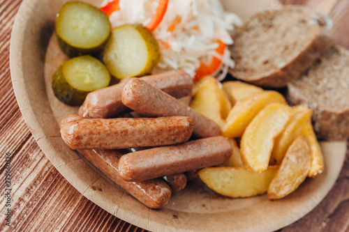 Tasty sausages with fried potatoes, cucumber, cabbage and chlobe in a paper plate on a wooden table.