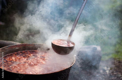 Outdoor rural summer scene where above the orange flames the traditional Hungarian goulash is boiling in a cauldron with a ladle