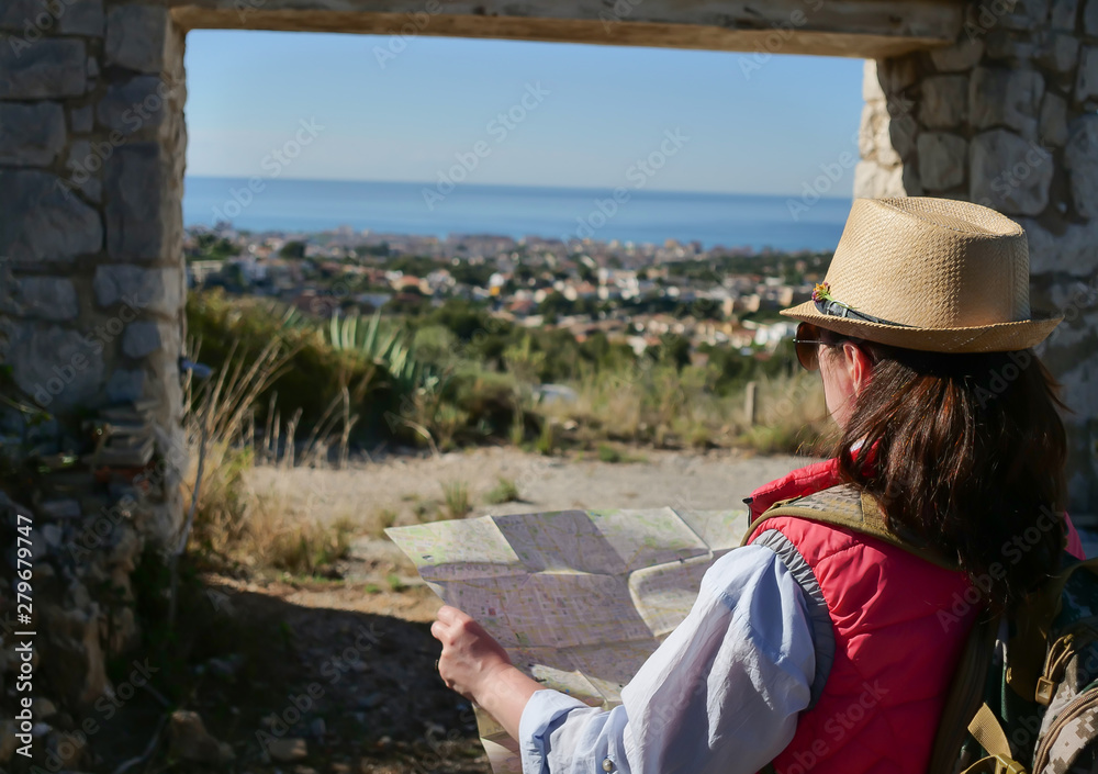 A girl tourist in a hat stands with a map of the area stands in front of a stone old gate, rear view