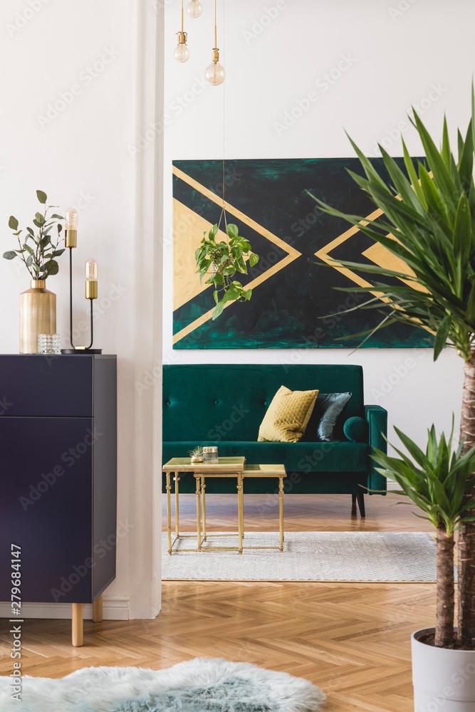 Fototapeta premium Modern and luxury interior of living room with shelf, gold coffee table velvet sofa, pillows, tropical plant and elegant accessories. Design paintings on the wall. Stylish home decor. Template