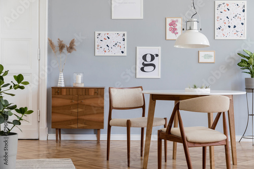 Stylish scandinavian home interior of sitting room with design wooden chairs, family table, plants, accessories and mock up posters gallery wall. Gray background walls. Modern home decor. Template.