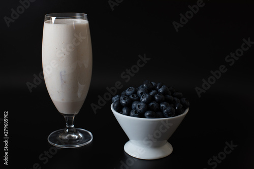 Blueberries in a white bowl and scattered around a glass of milkshake on a black background isolated