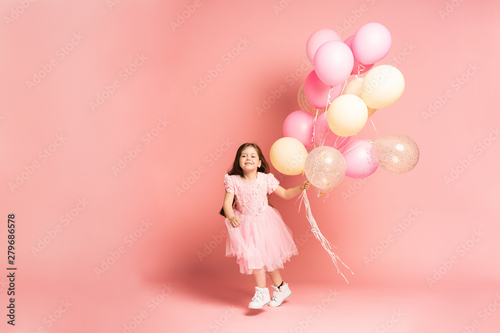 Happy celebration of birthday party with flying balloons of charming cute little girl in tulle dress smiling to camera isolated on pink background. Charming smile, expressing happiness Happy celebration of birthday party with flying balloons of charming cute little girl in tulle dress smiling to camera isolated on pink background. Charming smile, expressing happiness