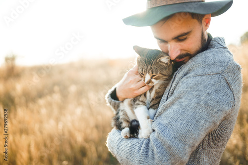 Lovely young hipster with a cat. A guy with a mustache and a beautiful smile is hugging a cat. Beautiful autumn sunset.