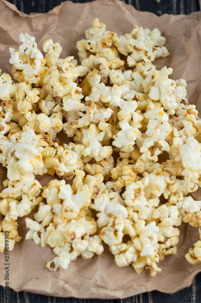 Homemade vegan fresh crunchy popcorn on brown craft paper lie on a dark wooden table. A useful snack for a movie, an idea for a meal. Dark mood background, top view. Homemade vegan fresh crunchy popcorn on brown craft paper lie on a dark wooden table. A useful snack for a movie, an idea for a meal. Dark mood background, top view.