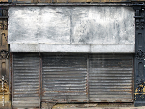the front of an abandoned store on a street with closed dirty rusting metal shutters over the shop front and door