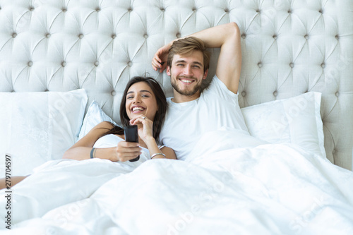 Photography happy couple with remote lying in bed at home and watching tv.